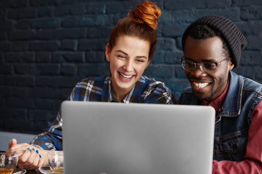 Imagem ilustrativa de uma mulher ruiva e um homem negro utilizando o notebook em um café.